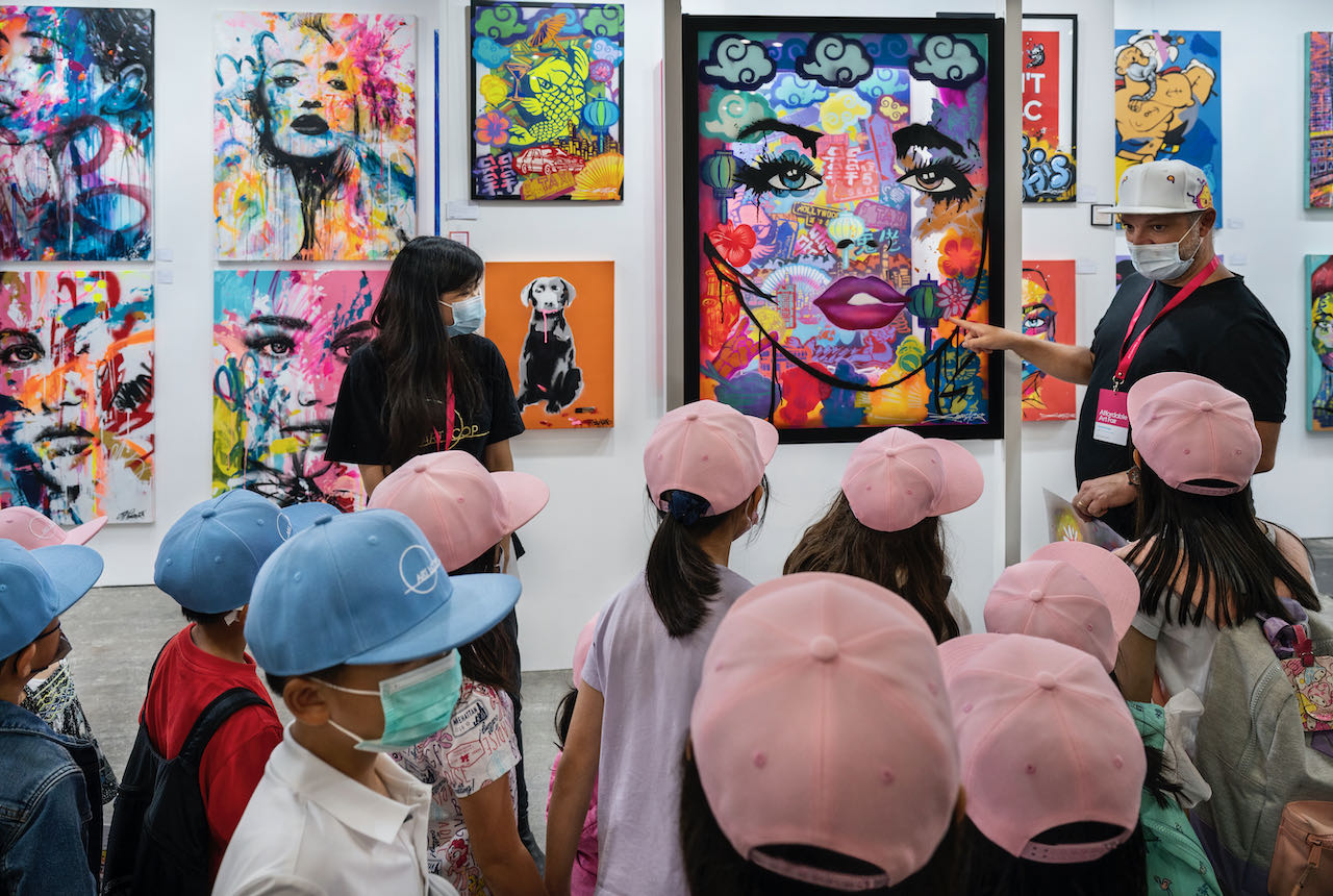 A group of children in pink and blue caps listening to tour guide introducing artworks on wall.