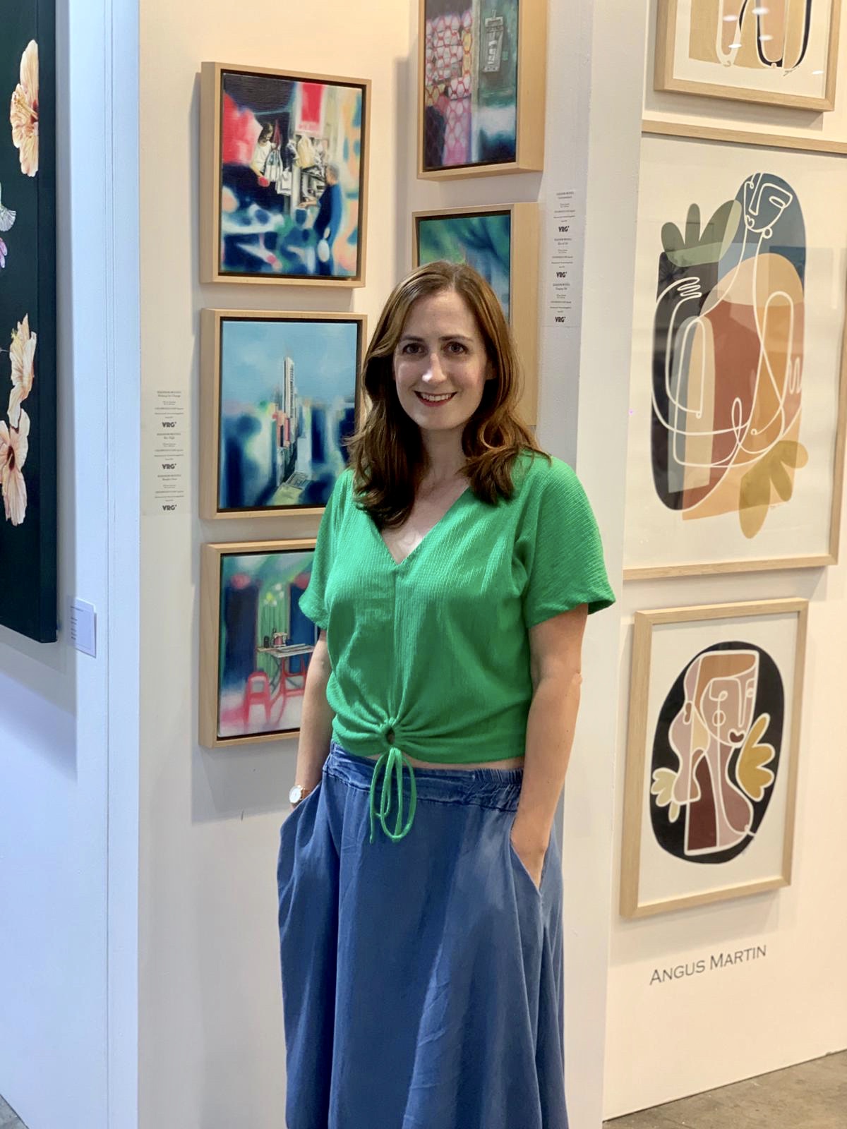 Eleanor standing in front of artworks, wearing a green shirts and smiling to the camera
