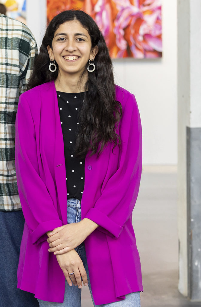 A portrait of Atena Abrahimia against the backdrop of Affordable Art Fair Amsterdam. She wears a bright fuchsia blazer and a spotted top.  