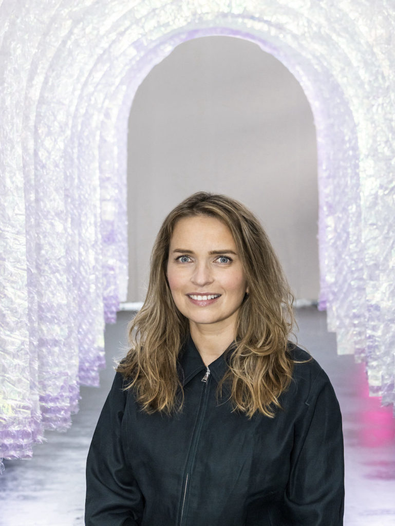 A portraits of Blythe Bolton stood underneath an art installation comprising of multiple clear plastic archways. She has blond, shoulder length hair and is smiling. She wears a black jacket zipped to the top. 