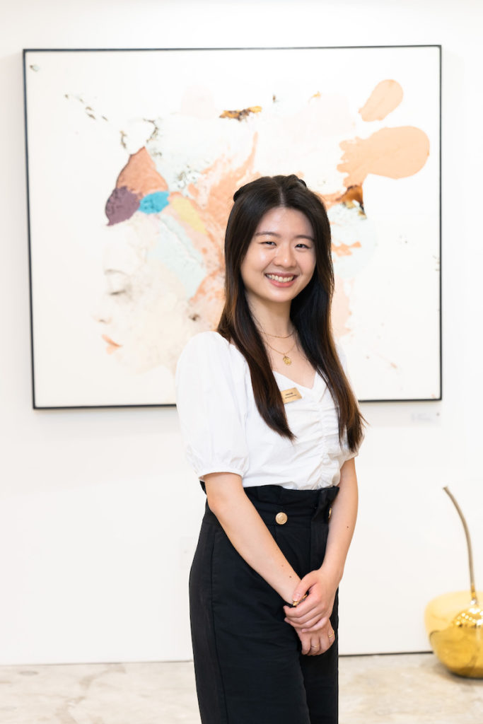 Coco Ma, Asia Marketing Manager, stands in front of an abstract artwork. She wears a white blouse and black trousers, with her hands clasped. 