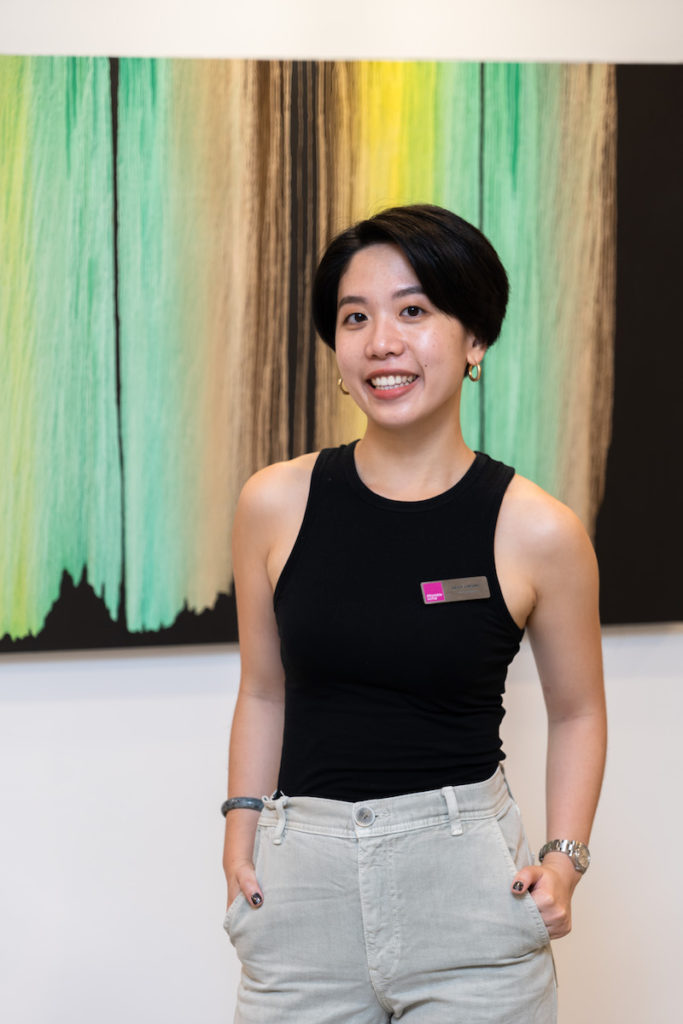 Kelly Cheung, Fair Coordinator, stands with her hands in her pockets in front of a painting. 