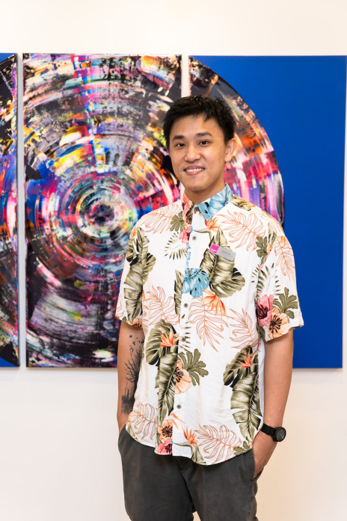 Anson Koh, Finance Co-ordinator for Asia, stands in front of an electric blue abstract artwork. He wears a floral shirt and grey trousers. 