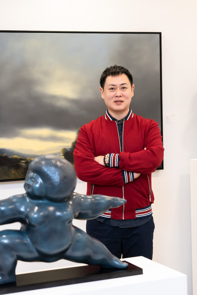 Fair Director Alan Koh stood next to a sculpture in a yoga pose, in front of a landscape painting with moody, grey clouds. 