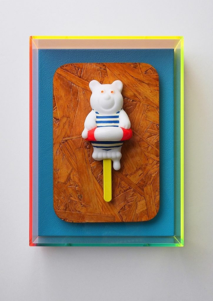 A cartoon polar bear lollipop mounted on wooden board, wearing a rubber ring and stripy swimsuit. The artwork is framed by perspex painted yellow, red, green and blue. 