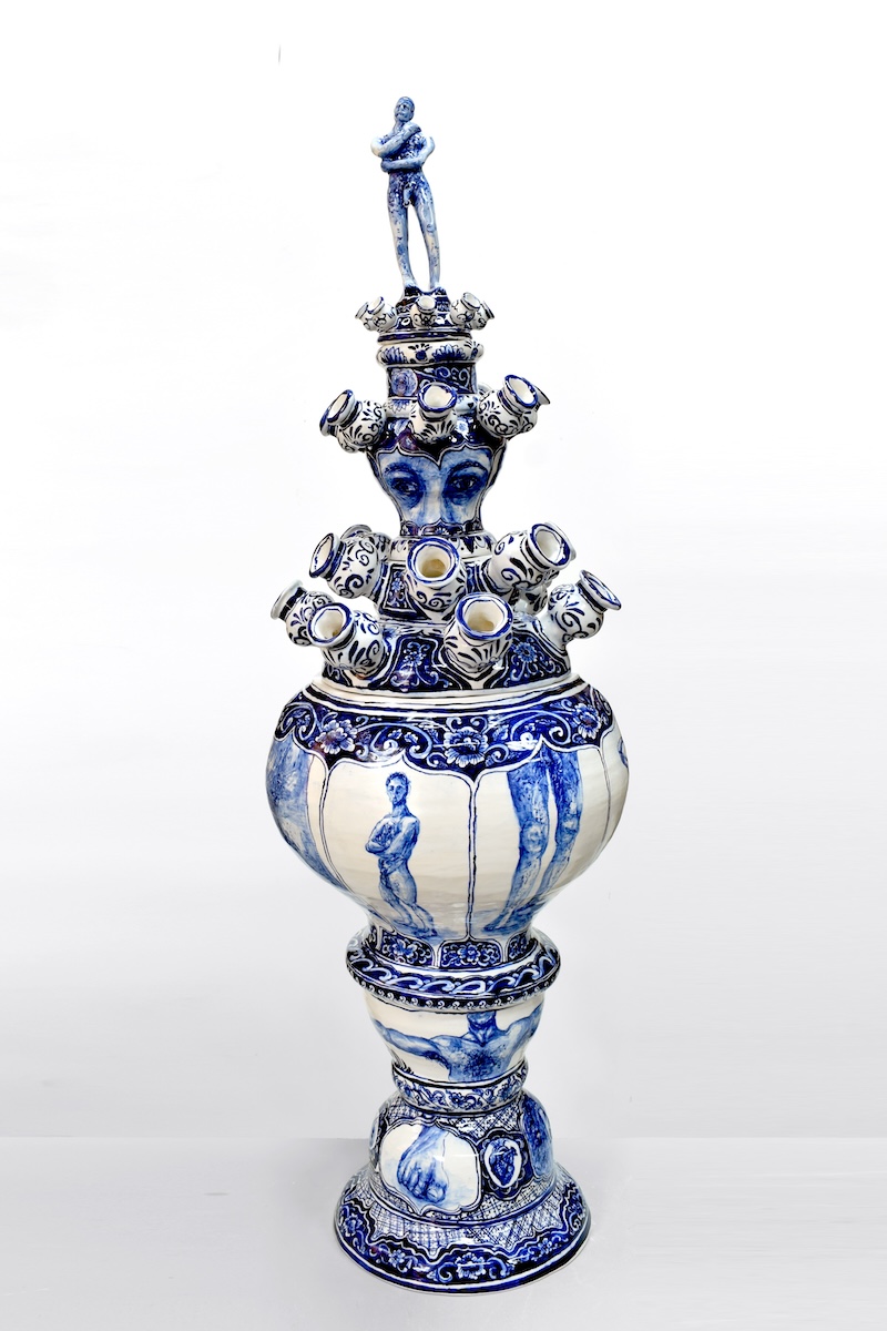 An intricate blue and white ceramic vase painted with male body parts and a male figure on top. 