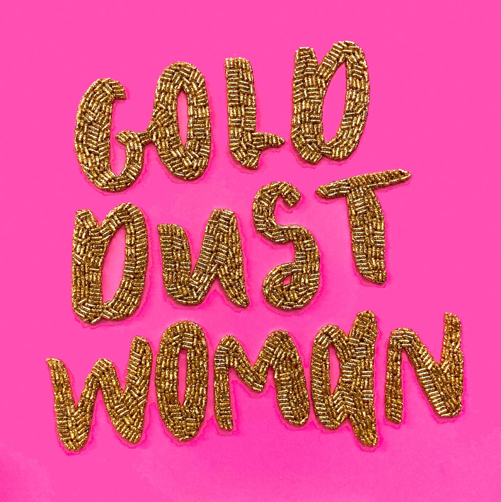 Gold beads spell out the words 'Gold Dust Woman' on a solid hot pink background. 