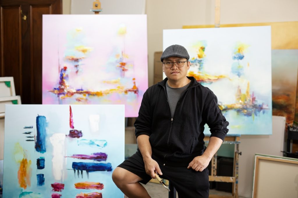 The male artist sitting among three of his abstract oil paintings, facing the camera and holding a palette knife in his right hand. 