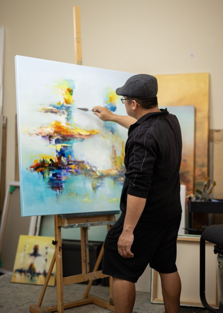 Male artist Keit Ancog painting with his right hand on a square-shape canvas, the colourful and abstract oil painting has a blue background and a mix of warm colours in the middle, ahead of Art Exhibition in Hong Kong. 