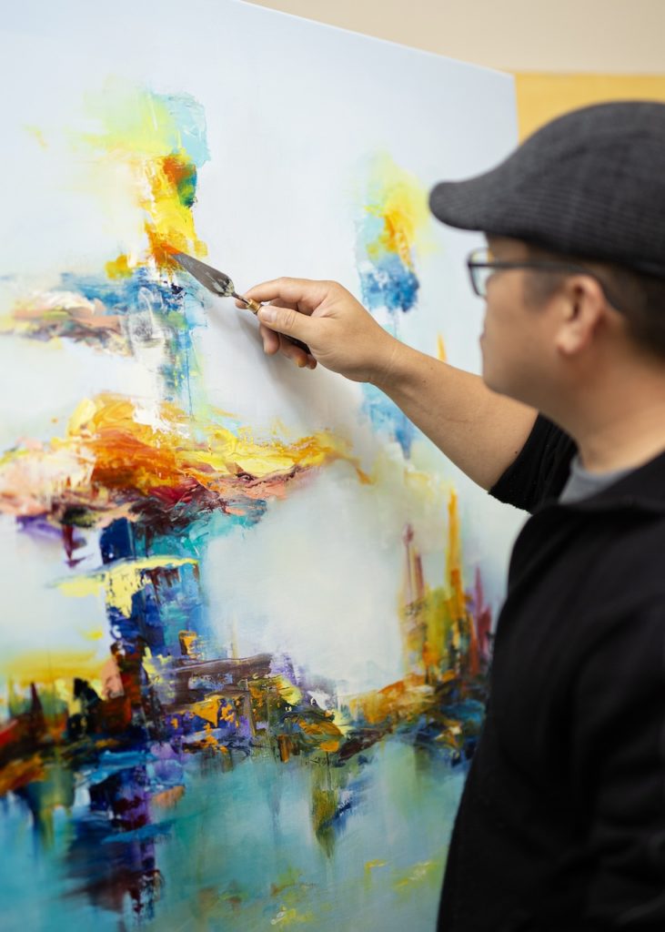 A male artist in a grey hat on the right, painting the oil canvas on the left side with his right hand. The oil painting is abstract, with a light blue background, and a colourful mix of warm colours forming thick and intersecting horizontal and vertical brush lines in the center. 